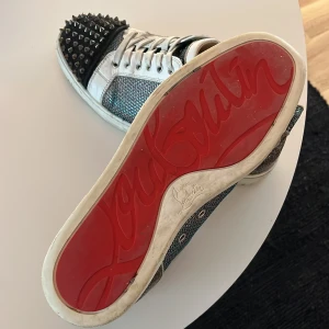 Christian Louboutin skor - Snygga sneakers med glittrigt material och nitar på tån. Skorna har en klassisk snörning och den ikoniska röda sulan.