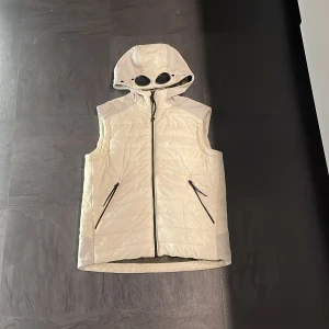 Vit  väst från C.P. Company - Säljer en stilren vit  väst från C.P. Company. Västen har en unik huva med inbyggda glasögon och dragkedja framtill. Perfekt för en cool och modern look. Köpt på NK. En liten reva fins på vänster sidan av luvan se bild.