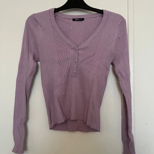 Lila ribbad tröja från Gina Tricot - Säljer en lila ribbad tröja från Gina Tricot med v-ringning och knappar framtill. Tröjan har långa ärmar och en bekväm passform. Passar även på M. Perfekt för en stilren look 💞