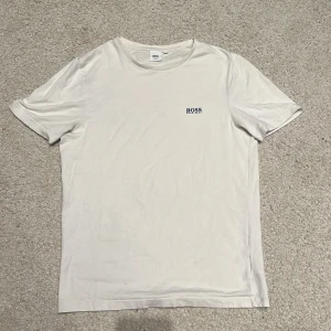 Vit t-shirt från Hugo Boss - Säljer en stilren vit t-shirt från Hugo Boss med blå logotyp på bröstet. T-shirten har en slim fit passform och är kortärmad. Perfekt för en minimalistisk look. Storlek S