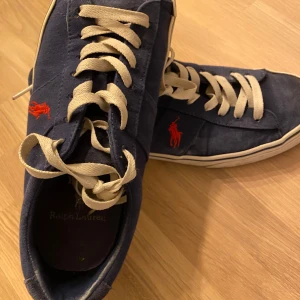 Blå sneakers från Ralph Lauren - Snygga blå sneakers från Ralph Lauren med vita snören och röd logga på sidan. Skorna har en klassisk design med en bekväm sula och är perfekta för en avslappnad stil.
