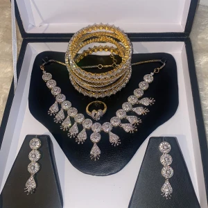 Glittrande smyckeset i guld och silver - Elegant smyckeset med ett halsband, örhängen, armband och ring. Smyckena är i guld och silver med glittrande stenar. Halsbandet och örhängena har droppformade detaljer, medan armbanden är staplade i olika mönster. Perfekt för att lysa upp vilken outfit som helst.