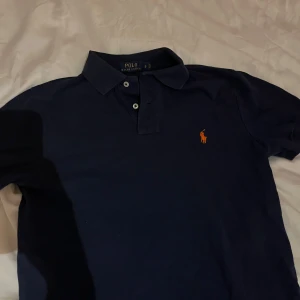 Mörkblå pikétröja från Ralph Lauren - Säljer en klassisk mörkblå pikétröja från Ralph Lauren med den ikoniska loggan i orange på bröstet. Tröjan har korta ärmar och knappar vid kragen. Perfekt för en stilren look.