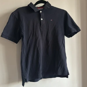 Mörkblå pikétröja från Tommy Hilfiger - Säljer en klassisk svart pikétröja från Tommy Hilfiger med knappar framtill och en liten logotyp på bröstet. Perfekt för en stilren look. Passar stl 152 