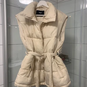Beige dunväst från Bik Bok - Dunväst från Bikbok med skärp i Storlek XS, perfekt till våren! Små fläckar som inte gått bort i tvätten tyvärr 