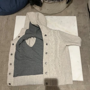 Beige stickad hoodie med knappar - Säljer en mysig beige stickad hoodie med knappar framtill. Tröjan har en kabelstickad design och ribbade muddar vid ärmslut och nederkant. Perfekt för kyliga dagar!