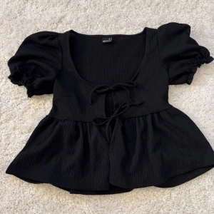 Svart topp från Gina Tricot - Snygg svart topp från Gina Tricot med puffiga kortärmar och knytning framtill. Toppen har en ribbad struktur och en lätt utsvängd nederdel som ger en fin siluett. Använd typ 1 gång!