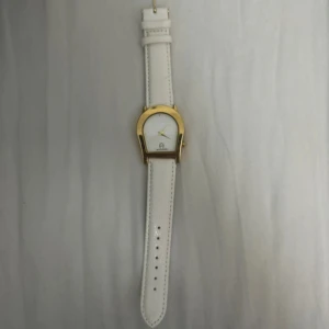 Vit klocka från Aigner med gulddetaljer - Elegant klocka från Aigner med vit läderrem och guldfärgad boett. Urtavlan är vit med subtila guldfärgade visare och Aigner-logotypen. Perfekt för den som söker en stilren och tidlös accessoar.
