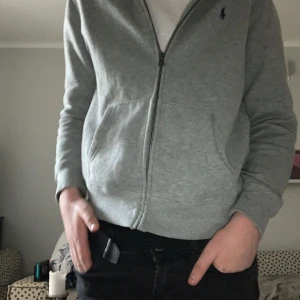Grå hoodie från Ralph Lauren - Säljer en stilren grå hoodie från Ralph Lauren med dragkedja och broderad logga på bröstet. Perfekt för en avslappnad look. Den har långa ärmar och en bekväm passform.