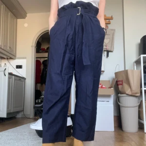 Mörkblå byxor med bälte från Arket - Snygga mörkblå byxor från Arket i ull. De har en hög midja och ett matchande bälte som ger en stilren look. Byxorna har en loose passform och är perfekta för en chic vardagsstil. Spännet på det inbyggda bältet har börjat tappa färg.