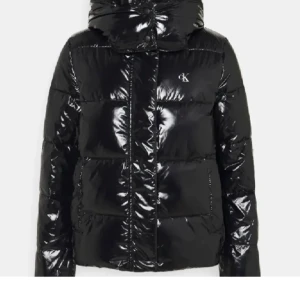 Svart glansig dunjacka från Calvin Klein - Snygg svart glansig dunjacka från Calvin Klein med huva och dragkedja. Jackan har en pufferdesign och CK-logga på bröstet. Köptes innan vintern nypris-ca 2100 beroende på sida. Storlek S