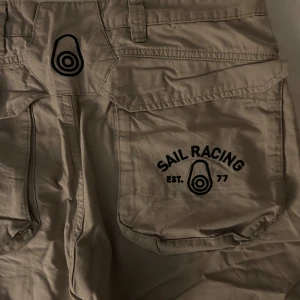 Beige shorts från Sail Racing - Snygga beige shorts från Sail Racing med praktiska fickor och cool logga. Perfekta för en avslappnad stil. De har en knapp och dragkedja framtill samt bälteshällor.
