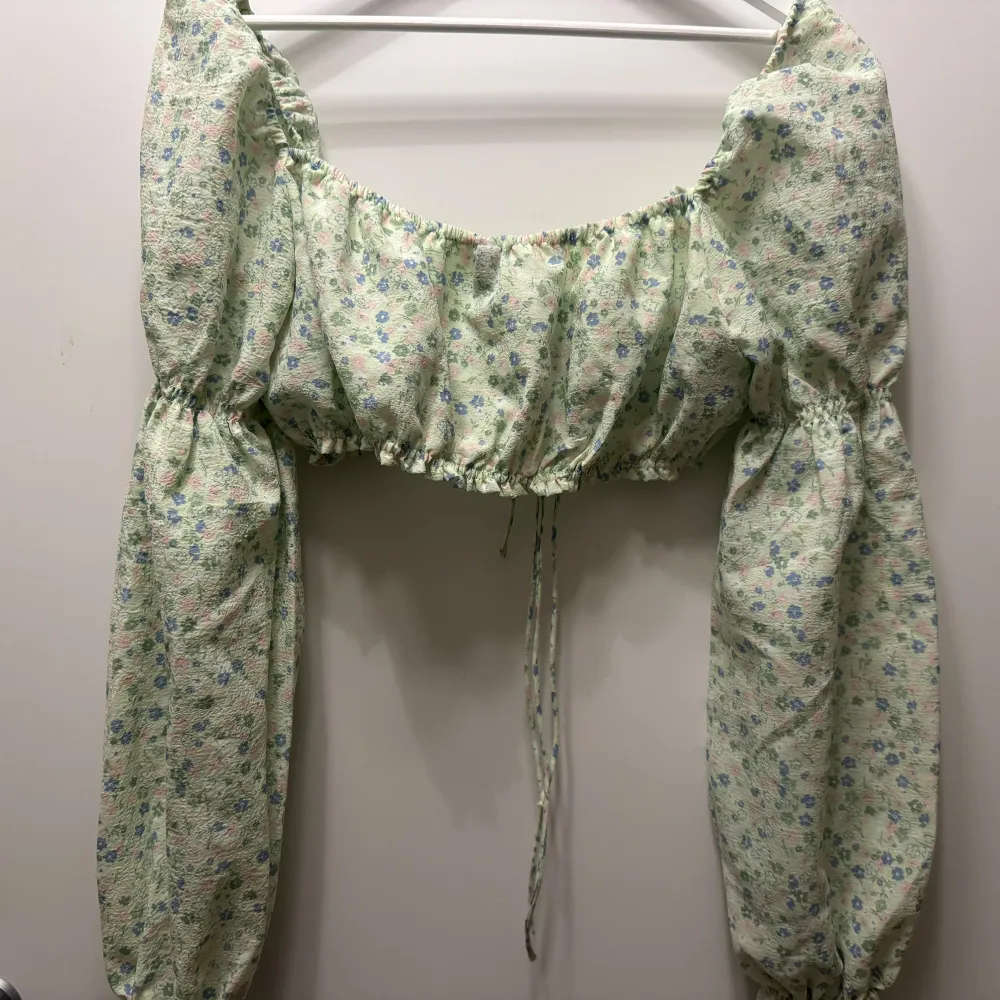 Blommig blus i ljusgrön med små blommor i rosa och blått. Blusen har långa ärmar och en söt knytning framtill. Perfekt för en somrig look 💖. Yläosat.