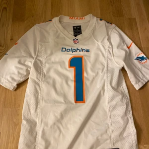 Vit Miami Dolphins fotbollströja - Säljer en vit Miami Dolphins fotbollströja från Nike med nummer 1 och texten 'Tagovailoa' på ryggen. Tröjan har korta ärmar och är dekorerad med blå och orange detaljer. Perfekt för fans av laget!
