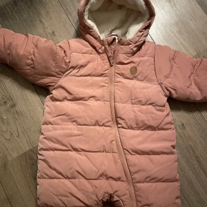 Rosa och grå babyoverall - Säljer två söta babyoveraller. Den första är en rosa pufferoverall med dragkedja och huva, perfekt för kyliga dagar. Den andra är en grå teddyoverall med dragkedja och huva, supermjuk och mysig för vintern.