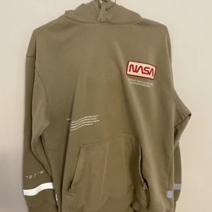NASA hoodie från H&M - Cool olivfärgad hoodie från H&M med NASA-logga på bröstet och amerikansk flagga på ärmen och reflex på nedre ryggen samt ärmar. Tryck 'National Aeronautics and Space Administration' finns på baksidan. Hoodien har en avslappnad passform, lite flagnat tryck (se sista bilden) övrig inga defekter!🌸