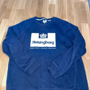 Mörkblå sweatshirt från Weekend Offender - Säljer en mörkblå sweatshirt från Weekend Offender med texten 'Helsingborg' och en krona på bröstet. Tröjan har långa ärmar och en rund halsringning. Perfekt för en avslappnad stil.