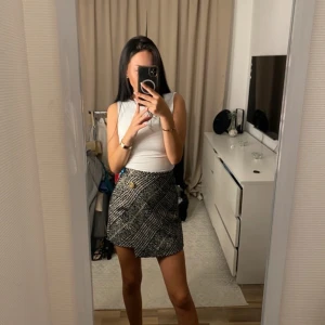 Rutig kortkjol med omlott - Snygg rutig kortkjol i svart och vit med omlott-design och en dekorativ knappdetalj framtill. Perfekt för en stilren look.