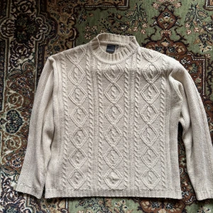 Beige stickad tröja från Oscars Basic - Säljer en stilren beige stickad tröja från Oscars Basic. Tröjan har ett snyggt flätmönster på framsidan och ribbade kanter vid hals, ärmslut och nederkant. Perfekt för kyliga dagar!