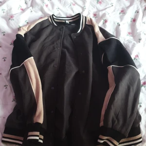 Svart och beige bomberjacka - Snygg svart bomberjacka med beige detaljer och randig krage. Jackan har knappar framtill och långa ärmar med ribbade muddar. Perfekt för en cool och avslappnad stil.