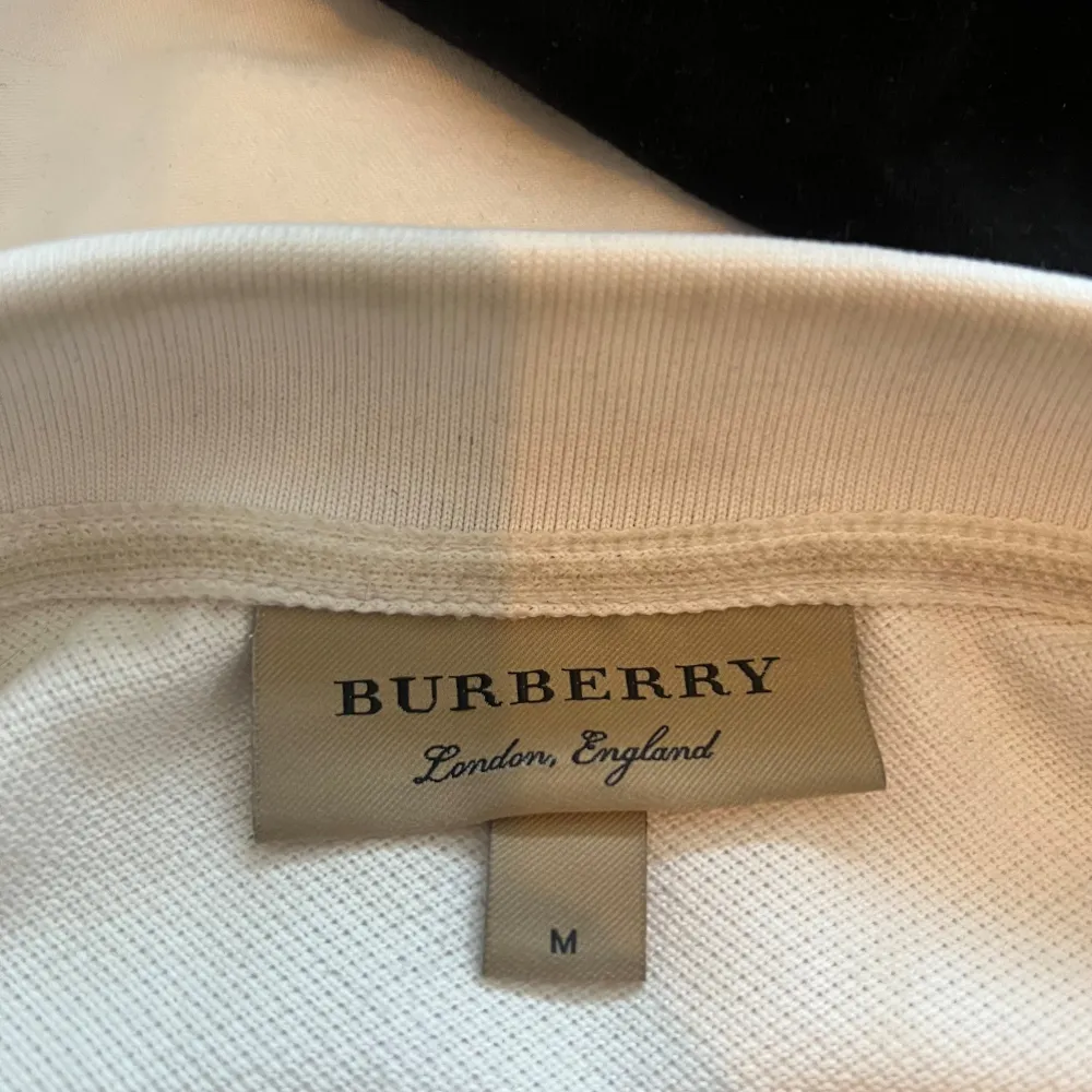 Säljer en stilren vit pikétröja från Burberry i storlek M. Tröjan har klassisk krage med knappar och ett diskret broderat märke på bröstet. Insidan av kragen har det ikoniska Burberry-mönstret. Perfekt för en elegant och avslappnad look.. Tröjor & Koftor.