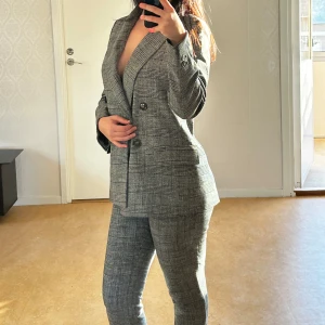 H&M - Kostym Set - Dam - Snygg grå rutig kostym i mycket bra skick. Kavajen har en dubbelknäppt design med svarta knappar och en klassisk passform. Byxorna är slim fit och matchar perfekt med kavajen. Perfekt för höst och vår, eller varför inte till en fest? Materialet känns som en blandning av ull och polyester, vilket ger en skön och stilren look.