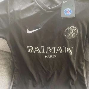Svart PSG x Balmain fotbollströja - Säljer en snygg svart PSG x Balmain fotbollströja i samarbete med Nike. Tröjan är i nytt skick med etikett kvar. Den har korta ärmar och ett stilrent tryck med Balmain Paris-loggan på framsidan. Perfekt för både match och vardag!