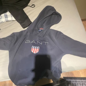 Mörkblå hoodie från GANT - Säljer en snygg mörkblå hoodie från GANT med deras klassiska logga och ett coolt emblem på bröstet. Perfekt för en avslappnad stil och passar bra till både jeans och joggers. Den har en stor och skön huva samt en praktisk magficka. Perfekt för höst och vinter!