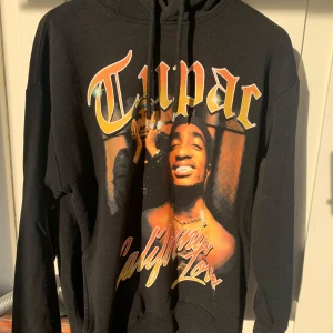 Svart Tupac hoodie från Pull&Bear - Säljer en svart hoodie från Pull&Bear med Tupac-tryck. Den har en stor bild av Tupac och texten 'California Love'. Perfekt för alla Tupac-fans. Mjuk och bekväm med fickor på varje sida och justerbar luva. Kanske något slitet på trycket annars i bra skick. ❤️❤️Priset går att diskutera. 