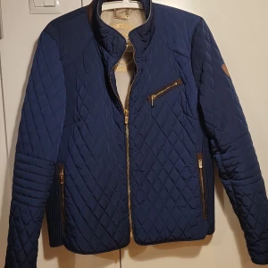 Blå quiltad jacka från Saki Sweden - Säljer en snygg blå quiltad jacka från Saki Sweden. Jackan har en dragkedja framtill och flera praktiska fickor med dragkedjor. Den är i mycket bra skick och perfekt för höst och vår. Stilig och bekväm med långa ärmar och fina detaljer som knappar vid ärmsluten.