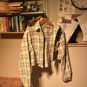 Rutig croppad fleece jacka från Urban Outfitters - Säljer en superfin rutig jacka från Urban Outfitters. Den är croppad och har en skön, avslappnad stil. Perfekt för höst och vår! Jackan har långa ärmar och knappar framtill. Färgerna är beige, blå och brun. Passar perfekt till en casual outfit.