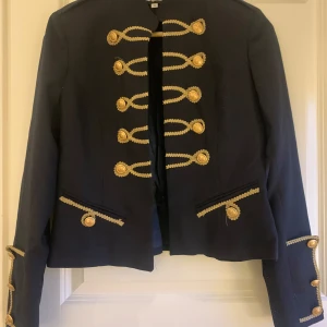 Marinblå kavaj med guldknappar från Rebel Cashmere - Säljer en elegant svart kavaj från Rebel Cashmere i storlek 36. Den har snygga guldknappar och dekorativa detaljer som ger en lyxig känsla. Perfekt för höst och vinter, och funkar både till fest och vardag. Kavajen är i mycket bra skick och redo för nya äventyr!