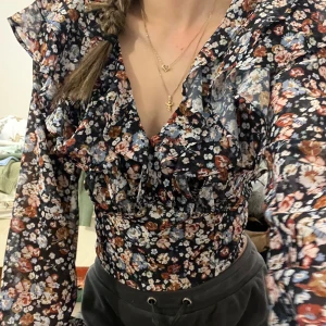Blommig blus med volangdetaljer - Säljer en superfin blommig blus med volanger och öppen rygg. Den har långa ärmar och ett vackert mönster i svart, vitt och brunt. Perfekt för både vardag och fest! 💐