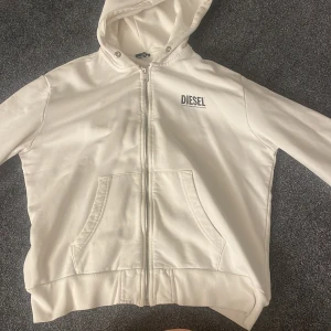 Vit hoodie från Diesel - Säljer en snygg vit hoodie från Diesel i mycket bra skick. Den har en dragkedja framtill och en stor logga på både bröstet och ryggen. Helt i nyskick då den inte kommit till använding, har inte bra koll va dessa hoodies säljs för men säljer min för 200kr, men kan gå ner till 150kr! 😊