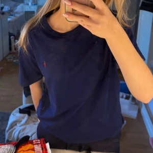 Mörkblå t-shirt från Ralph Lauren - Säljer en snygg mörkblå t-shirt från Ralph Lauren med det klassiska röda broderade logotypen på bröstet. T-shirten är i bra skick och perfekt för vardagsbruk. Den har en normal passform och korta ärmar. Perfekt för en avslappnad stil!