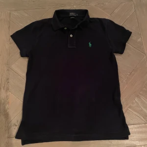 Svart pikétröja från Ralph Lauren - Säljer en klassisk svart pikétröja från Ralph Lauren i storlek S. Tröjan har en liten grön logga på bröstet och är i ett bekvämt bomullsmaterial. Perfekt för både vardag och lite finare tillfällen. Hör av dig vid intresse!