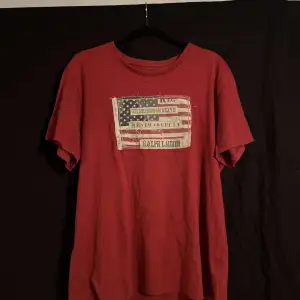 Säljer en röd t-shirt från Ralph Lauren med ett vintageinspirerat tryck av den amerikanska flaggan och texten 'Denim & Supply'. T-shirten är i 100% bomull och har en avslappnad passform. Perfekt för en casual look!