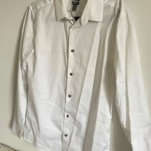 Vit skjorta från H&M - Säljer en klassisk vit skjorta från H&M i herrstorlek M. Skjortan är långärmad och har snygga svarta knappar. Perfekt för både vardag och fest! Den är i bra skick och gjord av bomull vilket gör den bekväm att bära. Passar perfekt till både jeans och kostymbyxor.
