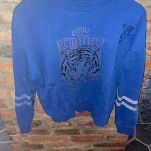 Blå sweatshirt från Gina Tricot - Cool blå sweatshirt från Gina Tricot med tryck av en tiger och texten 'Bronx New York'. Tröjan har slitna detaljer och vita ränder på ärmarna. Perfekt för en avslappnad stil med en edgy touch. Passar bra till jeans eller leggings! (Andvänt flitigt  + urtvättad färg därmed priset.)