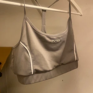 Grå sporttopp från DOM - Säljer en grå sporttopp från DOM i mycket bra skick. Toppen har en stilren design med vita detaljer och korsade axelband för extra stöd. Perfekt för träning eller en aktiv vardag. Materialet är mjukt och stretchigt för bästa komfort.