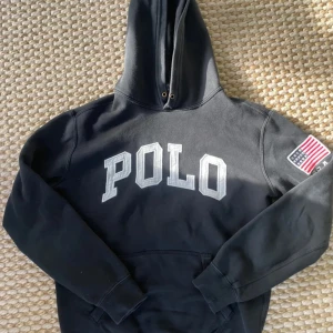 Svart hoodie från Ralph Lauren - Säljer en svart hoodie från Ralph Lauren med stor POLO-text på framsidan och en amerikansk flagga på ärmen. Perfekt för en avslappnad stil och passar bra till både jeans och joggers. Den är i mycket bra skick och superbekväm att ha på sig. Hör av dig om du är intresserad! 🖤
