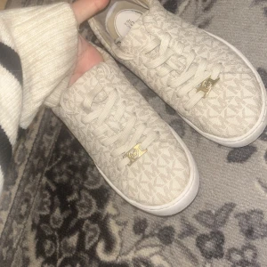 Beige sneakers med monogrammönster - Säljer ett par snygga beige sneakers från Michael Kors i mycket bra skick. De har ett diskret monogrammönster och guldfärgade detaljer vid snörningen. Perfekta för både vardag och festliga tillfällen. Storlek dam 36 passar mig som har 37 också 