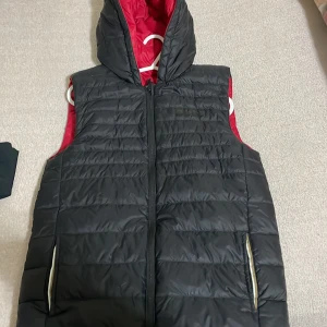 Svart och röd vändbar dunväst - Säljer en snygg vändbar dunväst i svart och rött. Västen har en dragkedja framtill och en praktisk huva. Perfekt för höst och vår när du vill ha lite extra värme utan att bära en jacka. Den är i mycket bra skick och passar till både vardag och träning.