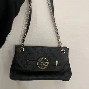 Svart axelväska med kedjedetaljer - Snygg svart axelväska med kedjedetaljer och ett stort metallmärke på framsidan. Väskan har ett elegant mönster och en praktisk dragkedja inuti. Perfekt för både vardag och fest!