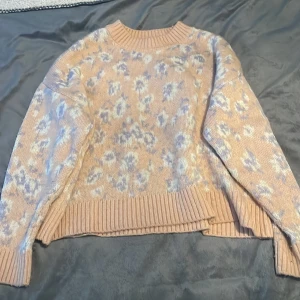 Rosa stickad tröja från Mango - Superfin rosa stickad tröja från Mango med ett subtilt blommönster i vitt och ljuslila. Den har en ribbad kant vid ärmslut och nederkant, vilket ger en snygg finish. Perfekt för kyliga dagar när du vill vara både varm och stilren. 