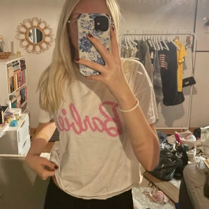 Vit t-shirt med barbie tryck - Säljer en vit t-shirt med rosa text 'Barbie' på framsidan. Den är kortärmad och har en avslappnad passform, perfekt för en casual look. T-shirten är i bra skick och passar till vardags eller en chill dag hemma.