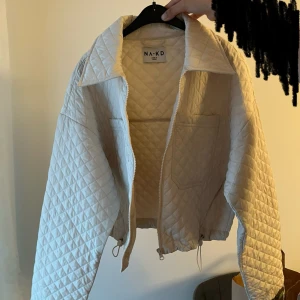 Beige quiltad jacka från NA-KD - Säljer en superfin beige quiltad jacka från NA-KD. Den har en dragkedja framtill och två stora fickor på bröstet. Jackan är perfekt för höst och vår, och ger en stilren look med sin krage och quiltade mönster. Den är i nyskick och passar perfekt för dig som vill ha något både snyggt och bekvämt. Jackan är oversized och passar i princip alla storlekar mellan S-L.