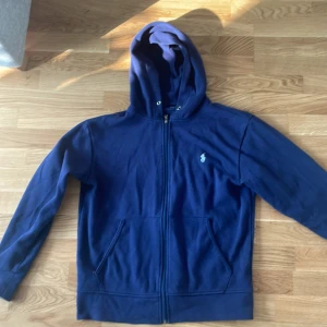 Mörkblå hoodie från Ralph Lauren - Säljer en snygg mörkblå hoodie från Ralph Lauren. Den har en dragkedja framtill och den klassiska broderade loggan på bröstet. Perfekt för höst och vinter, och den är i bra skick. Passar både till vardags och chill dagar. Hör av dig om du är intresserad! 💙
