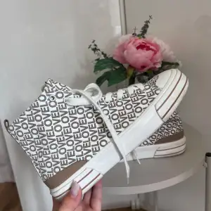 Säljer ett par coola vita sneakers från Graceland med ett unikt textmönster där det står 'LOVE' över hela skon. De har en brun detalj runt hälen och en vit sula med bruna ränder. Skorna har snörning framtill och är så gott som nyskick. Perfekta för vår och sommar!