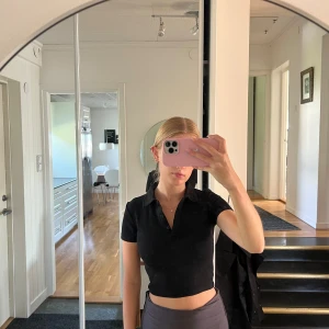 Svart croppad polotopp - Säljer en snygg svart croppad polotopp med korta ärmar. Perfekt för en casual look eller att klä upp för fest. Toppen har en tight passform och är superbekväm.